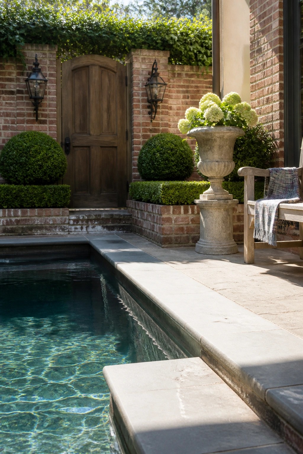 A rectangular pool with stone coping sits next to a brick patio enclosed by walls, a wooden door, topiary shrubs, and a large stone urn planter.