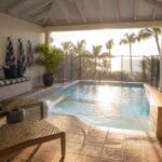 A covered pool area with turquoise water, a built-in bench with striped cushions, wicker lounge chairs on terracotta tile, and an ocean view beyond a glass railing.