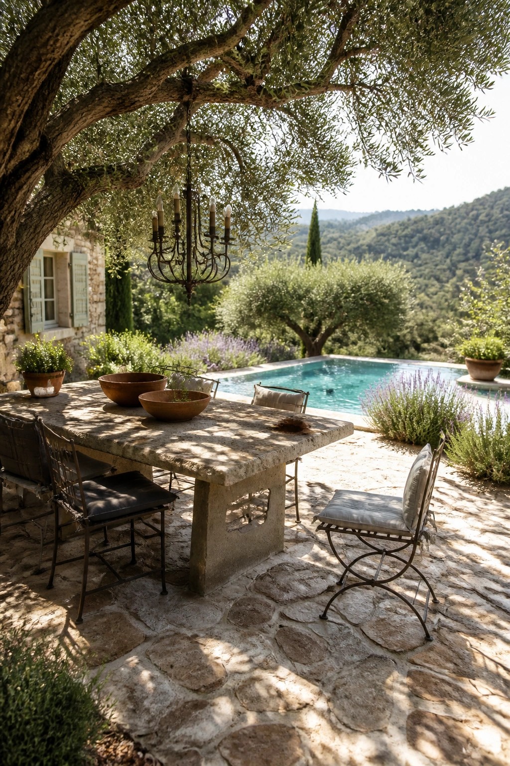 20 Practical Poolside Shade Ideas for Seating, Dining, and Relaxation Areas A stone dining table with metal chairs is positioned under a large tree with a chandelier hanging from its branches, next to a swimming pool on a stone patio.