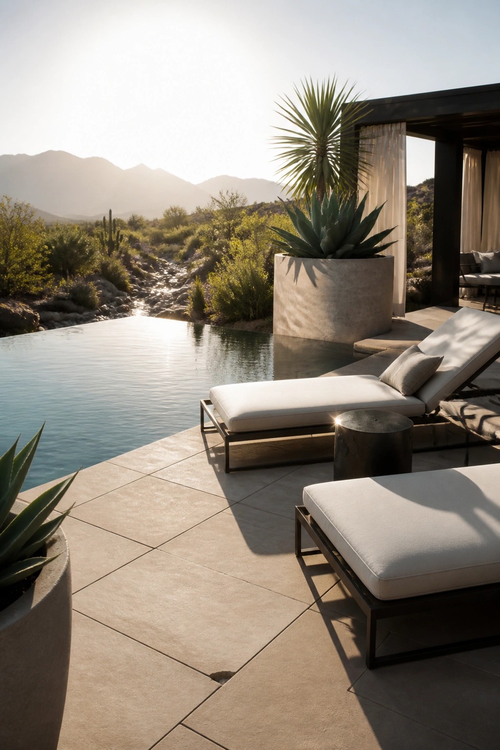 Two lounge chairs with white cushions sit on a tiled patio next to an infinity pool, with large concrete planters holding agave plants and a desert landscape in the background.
