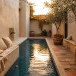 A narrow rectangular pool with blue mosaic tiles runs beside a stucco wall with a brass spout, opposite a built-in bench with beige cushions, potted trees, and string lights overhead.