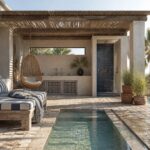 An outdoor poolside space with a wooden slatted ceiling, hanging wicker chair, built-in daybed, blue tiled shower, and potted plants on a stone floor next to a swimming pool.