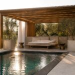 A wooden pergola with a slatted roof shelters a long cushioned bench beside a rectangular pool, flanked by potted olive trees and white curtains.