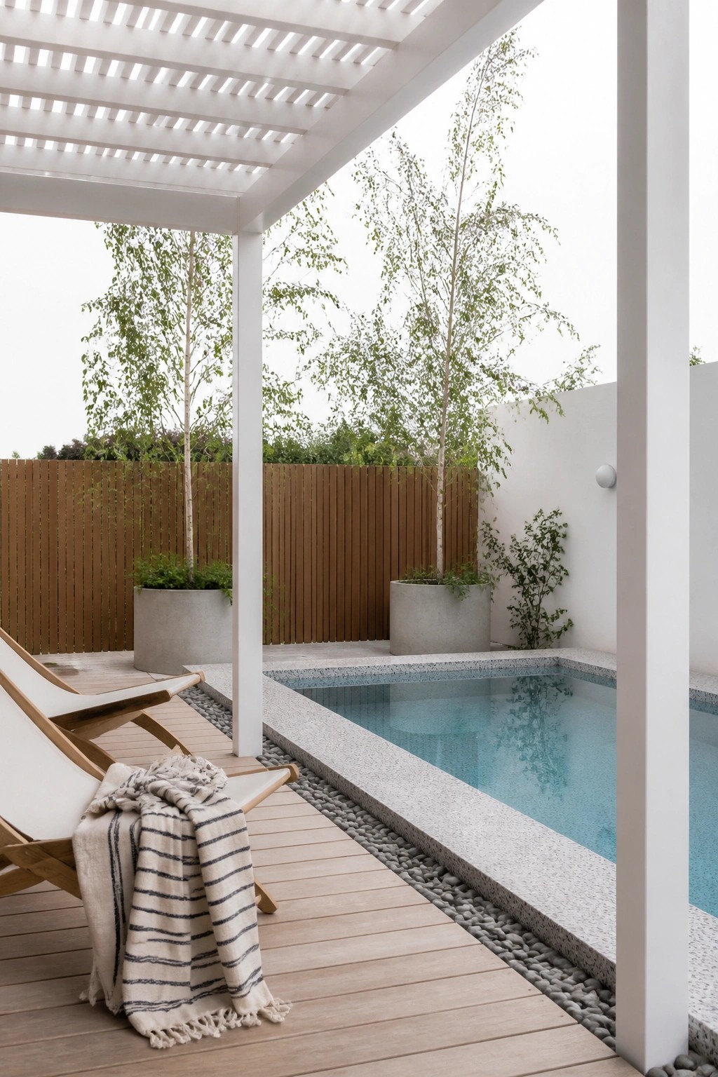 Modern patio with wooden lounge chairs and blanket beside pool under white pergola
