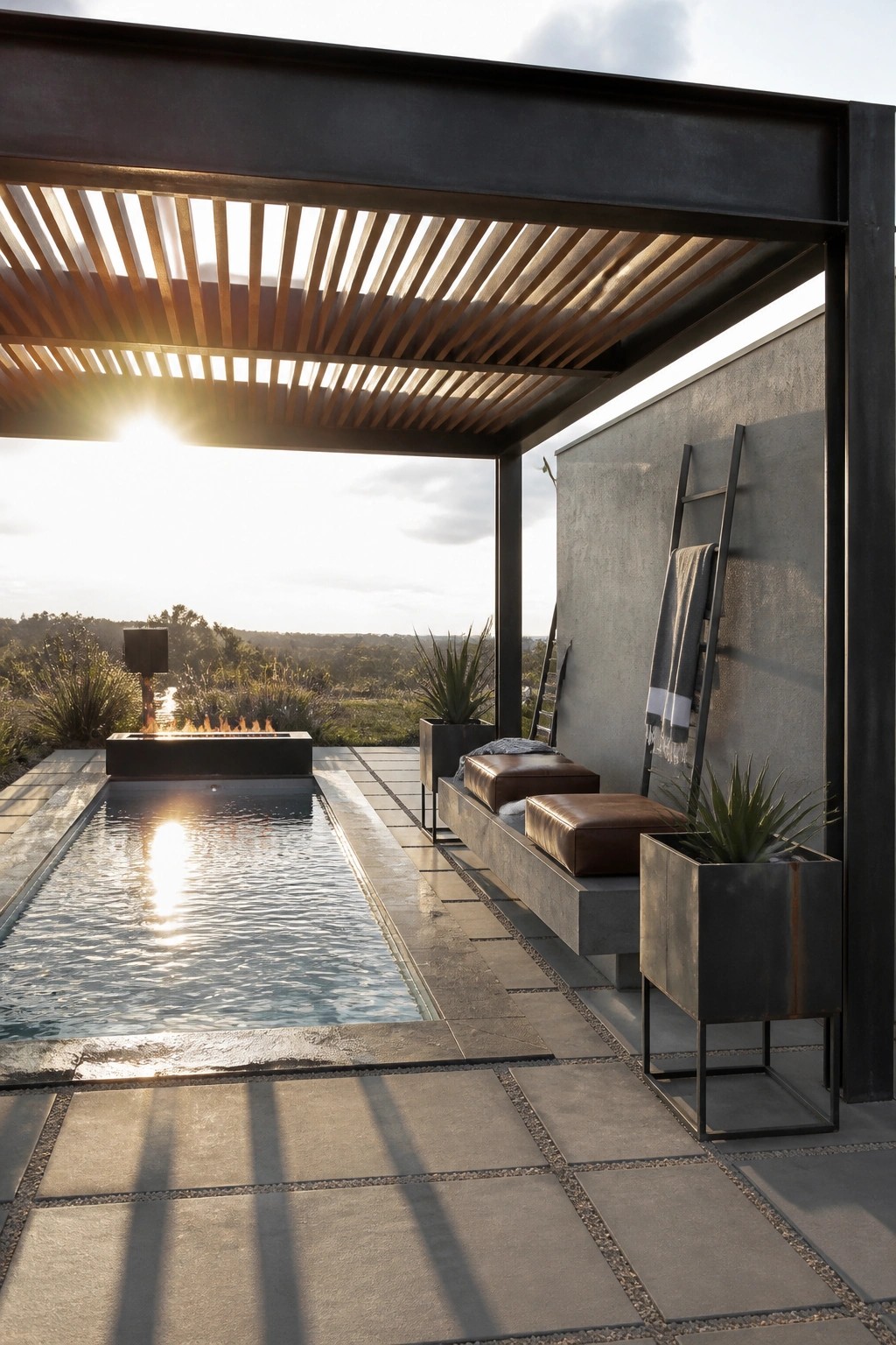 Modern outdoor pool area with a dark metal pergola featuring horizontal wooden slats, a long concrete bench with brown leather cushions, potted plants, and a fire pit beside the water at sunset.