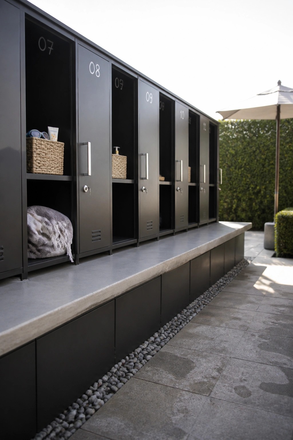 A row of black metal lockers numbered 07 through 11 with some doors open to show baskets and towels, built along a concrete patio bench with a pebble border.