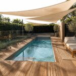 A long rectangular pool with turquoise water is covered by a large beige fabric shade sail stretched overhead, with wooden decking and lounge chairs along one side.