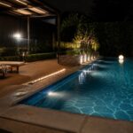 A nighttime view of a modern rectangular pool with a linear fire feature built into the coping, wooden deck with two lounge chairs, and a glowing white sphere floating in the water.