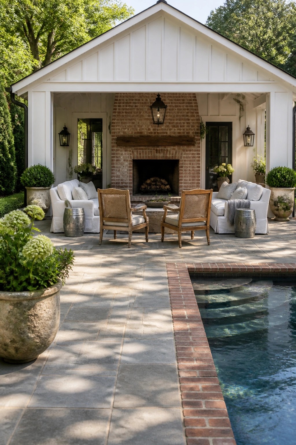 A white gabled cabana with brick fireplace, two sofas and two cane chairs on a stone patio next to a swimming pool.