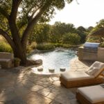 A freeform pool curves around a large tree with a stone patio, lounge chairs, and an outdoor kitchen area.