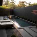 A long narrow pool runs beside a gravel path lined with rectangular stepping stones, with two lounge chairs at the far end and a large planter holding a small tree against a dark wall.