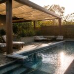 A wooden pergola with light fabric canopy shades striped lounge chairs next to a rectangular swimming pool with tiled steps and a metal handrail.