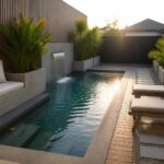 A long narrow pool with dark water runs alongside a wooden deck, with built-in concrete seating and cushions on the left and a small waterfall at the far end.