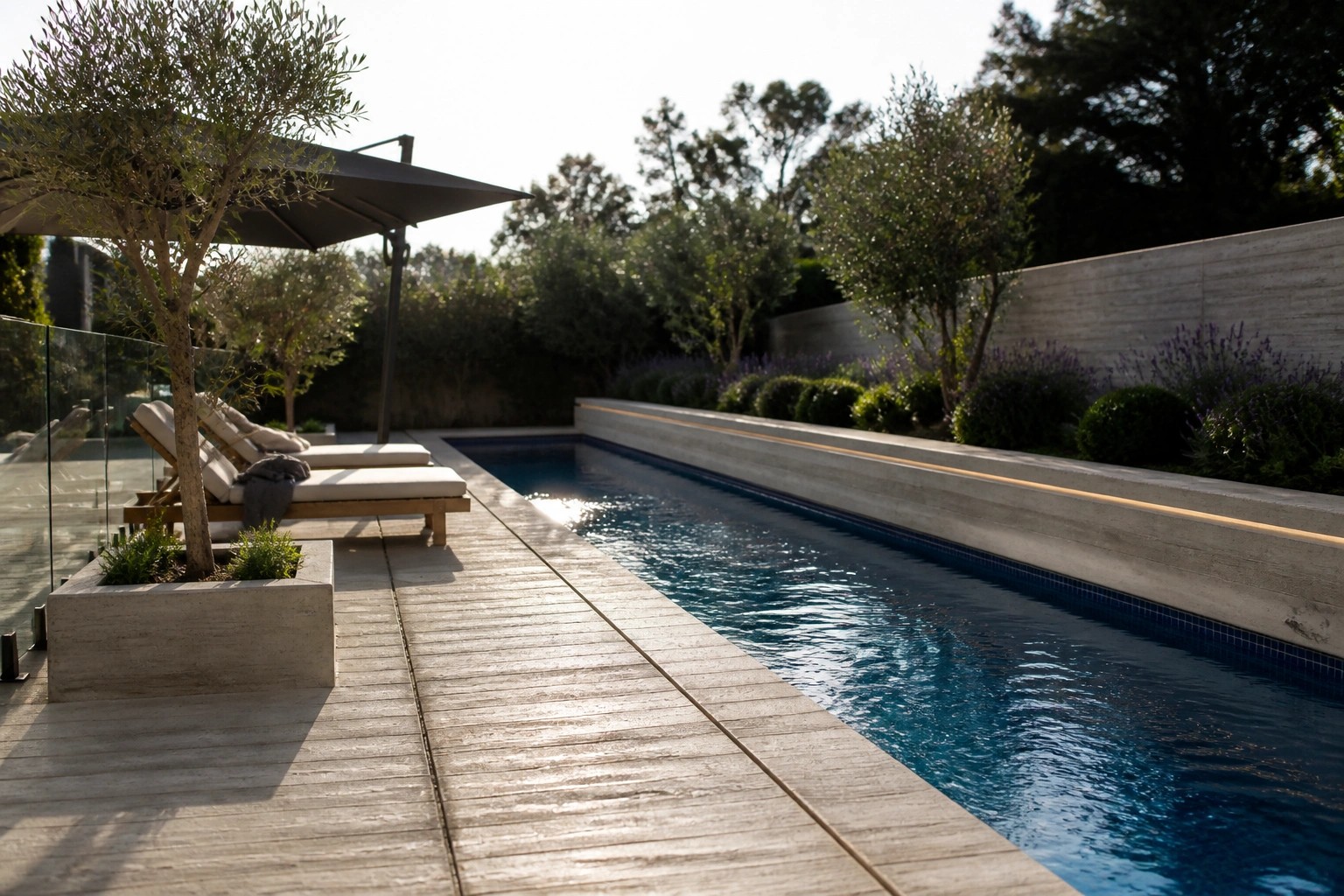 A rectangular pool bordered by wide concrete coping, with a wooden deck path, gray lounge chairs under a black umbrella, and concrete planters with round shrubs.