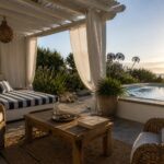 A covered poolside seating area with a striped daybed, wicker chairs, a wooden coffee table, hanging white curtains, and a view of the pool and ocean beyond.