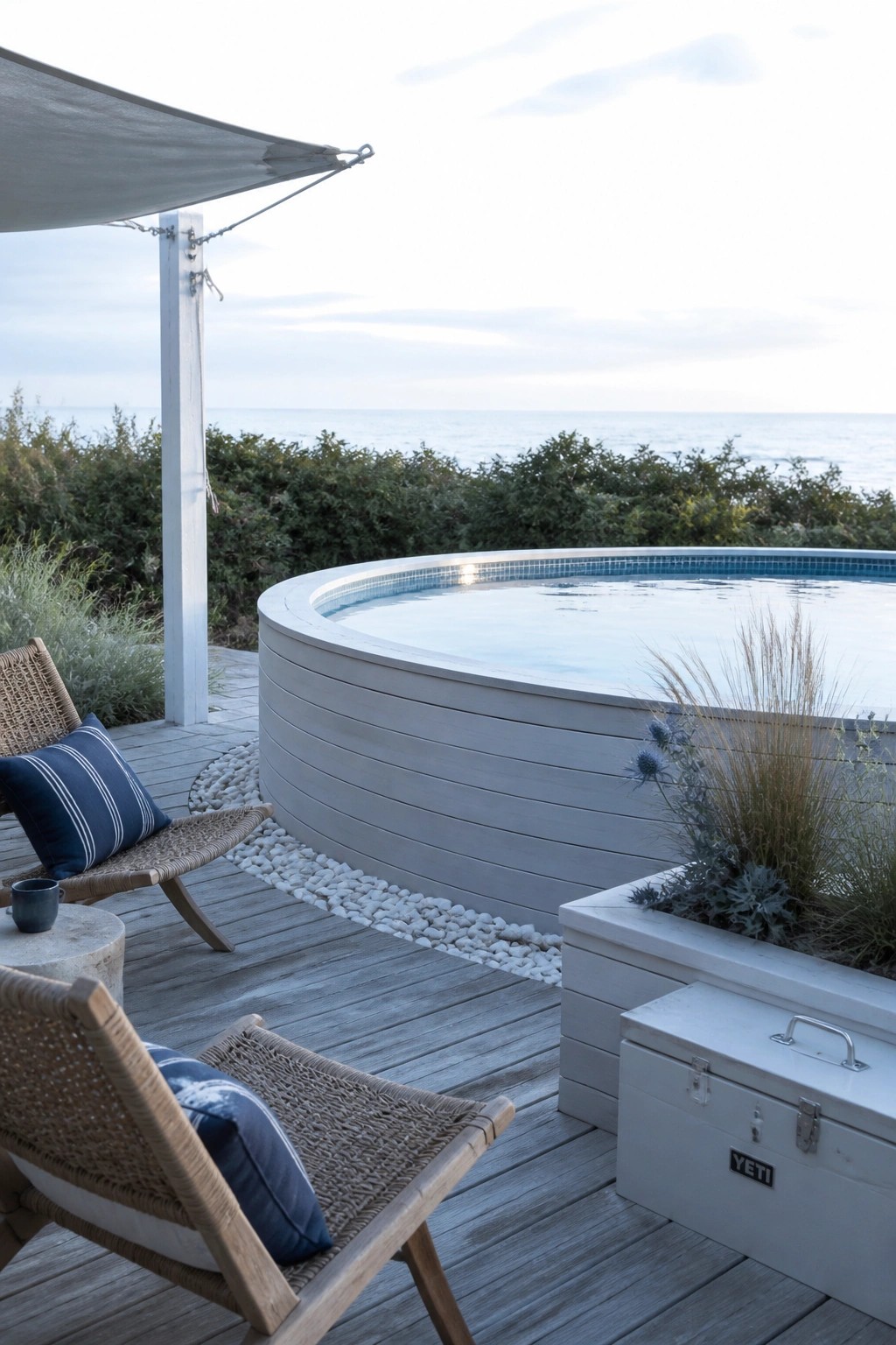23 Clever Above Ground Pool Patio Ideas That Feel Custom and Well Integrated An above-ground pool with light gray wood surround sits on a wooden deck overlooking the ocean, with woven chairs, a white planter box, and a white storage chest nearby.