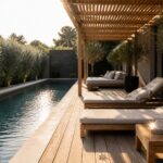 A long swimming pool beside a wooden deck with beige lounge chairs positioned under a slatted wooden pergola and potted trees along the edge.