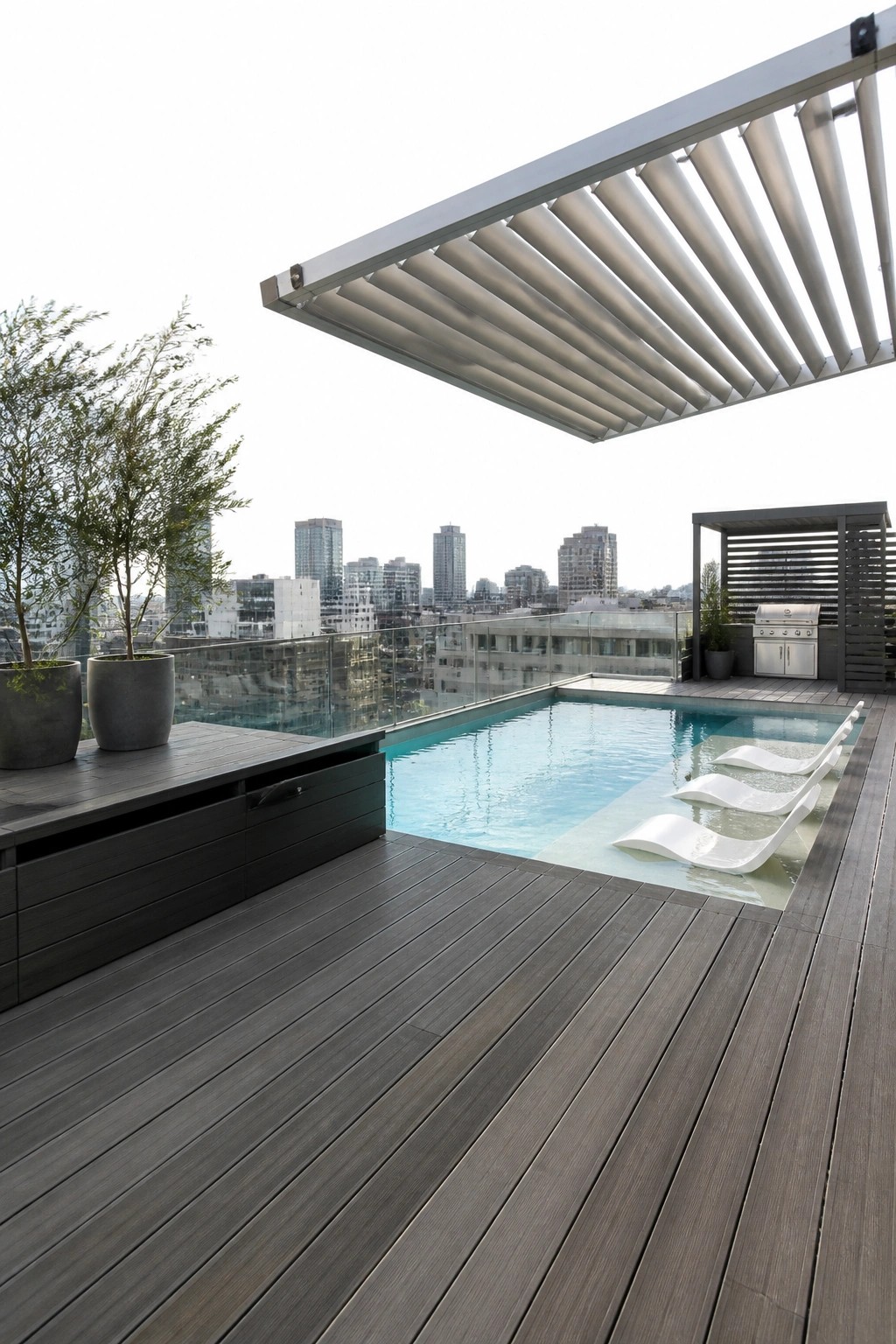 22 Contemporary Pool Shade Ideas in Minimalist Outdoor Settings A modern rooftop pool area with a large white louvered shade structure, dark wood decking, white in-pool lounge chairs, and potted trees overlooking a city skyline.