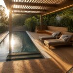 A rectangular swimming pool with blue mosaic tiles borders a wooden deck that holds two gray lounge chairs beneath a slatted wooden pergola structure.