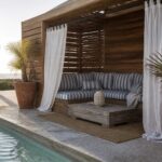 A wooden pool cabana with a daybed, striped cushions, white curtains on a rod, and large terracotta pots next to a swimming pool.