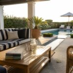 A wooden outdoor sectional with blue and white striped cushions sits on a stone patio beside a long swimming pool, with a matching striped umbrella and two wicker chairs nearby.