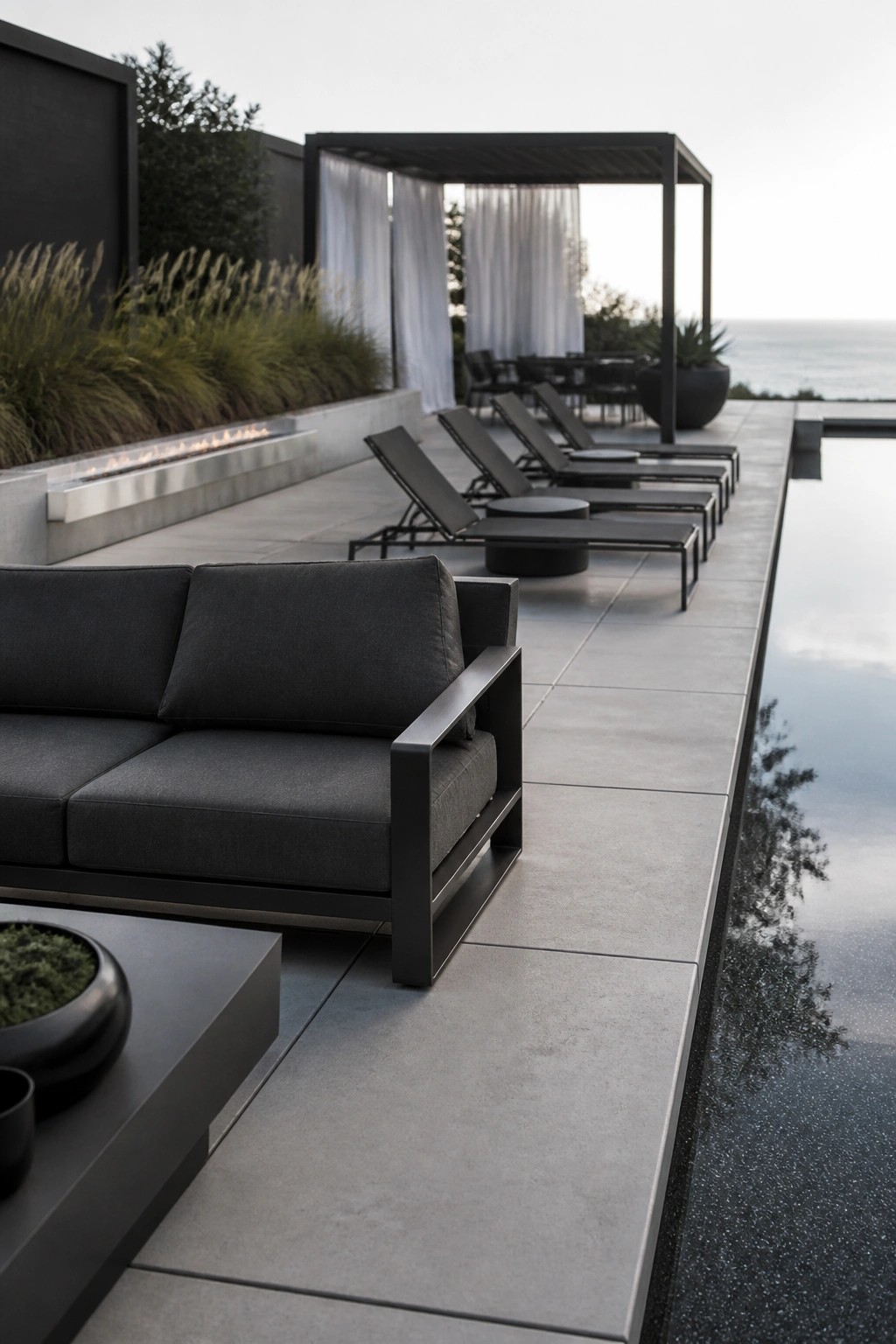 23 Fresh Pool Patio Furniture Ideas That Create Comfortable Outdoor Gathering Spaces Dark sofa and lounge chairs on concrete patio by infinity pool overlooking ocean