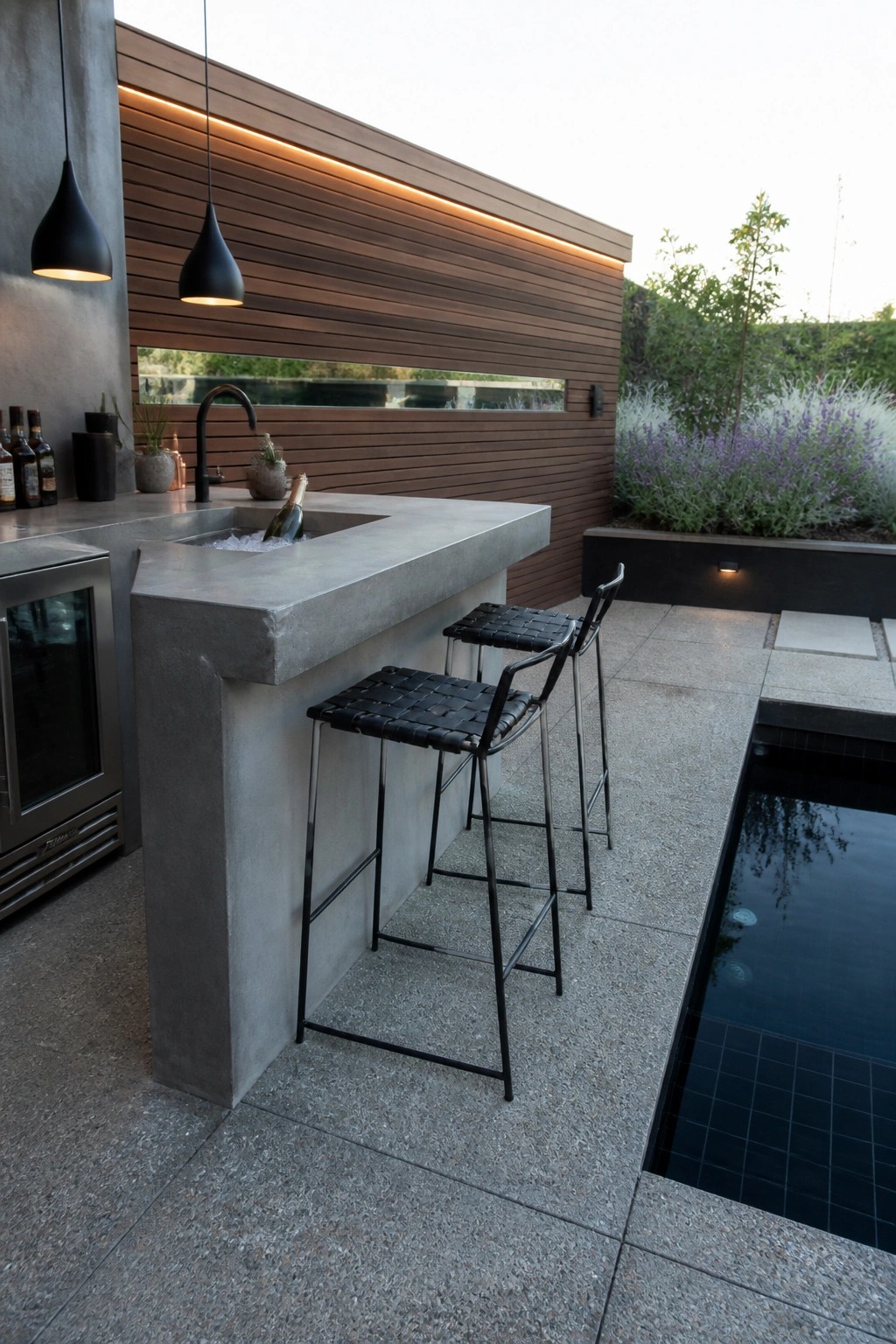 An outdoor concrete bar with a sink and two black woven stools sits on a stone patio beside a swimming pool.