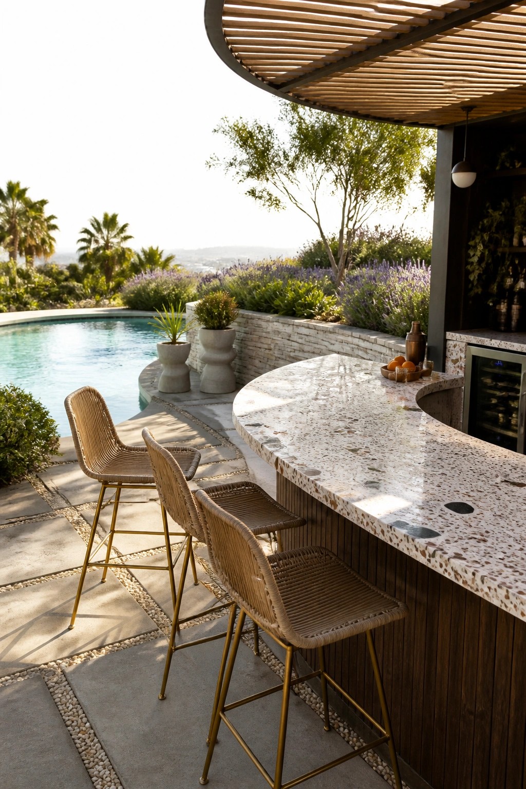 An outdoor curved bar with a speckled stone countertop and woven bar stools sits beside a swimming pool with surrounding plants and trees.