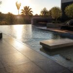 Modern outdoor pool with iridescent mosaic tile edging, large gray concrete deck tiles, dark lounge chairs, and a built-in fire feature in the background.