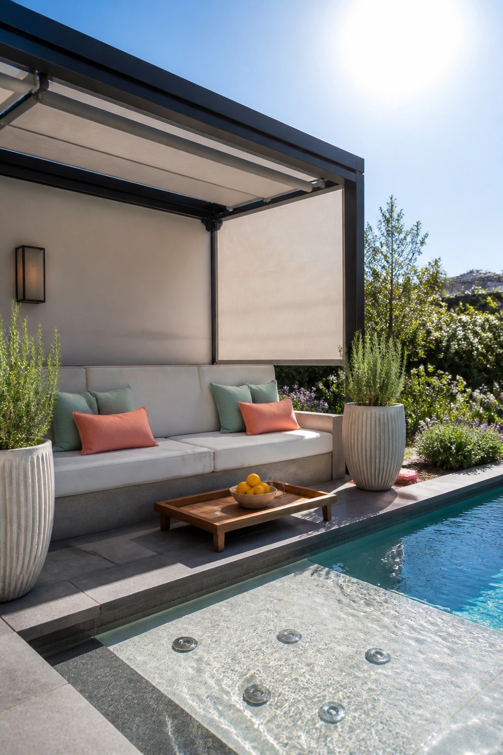 A modern black pergola with a beige drop-down side shade beside a swimming pool, showing built-in cushioned seating and two large potted plants.