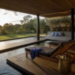 A covered wooden pool deck with lounge chairs, a side bench with pillows, and a long rectangular pool extending toward trees.