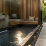 A narrow rectangular pool with dark tile, a waterfall spout on the left wall, and a long wooden bench built into the right edge, surrounded by wood fencing and pebble borders.