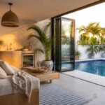 An interior view of a pool house showing a bed with beige linens, a small kitchenette, and open glass doors leading directly to a swimming pool with palm trees visible outside.