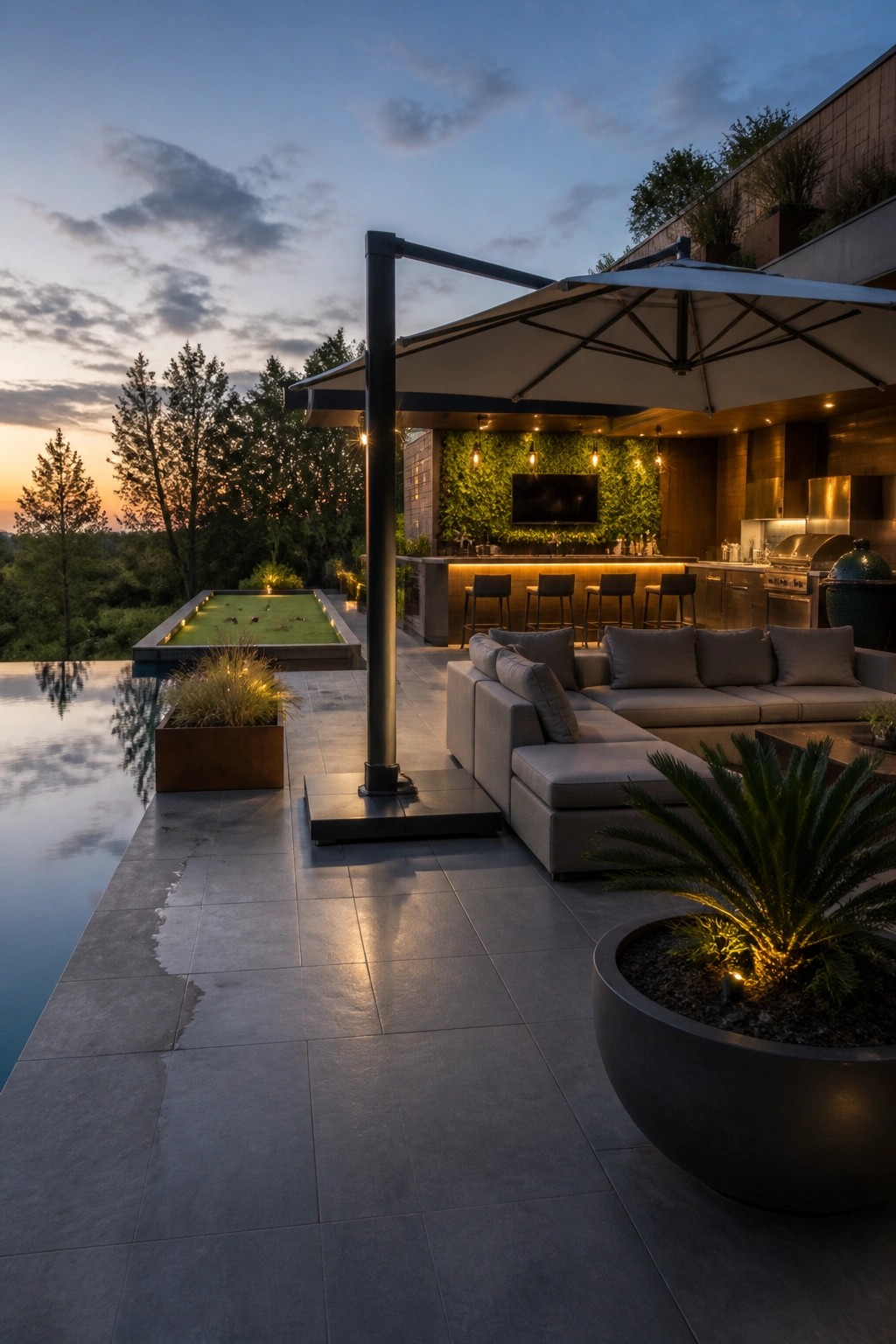 Modern backyard patio with large gray sectional sofa under a white cantilever umbrella next to an infinity pool and outdoor bar at dusk.