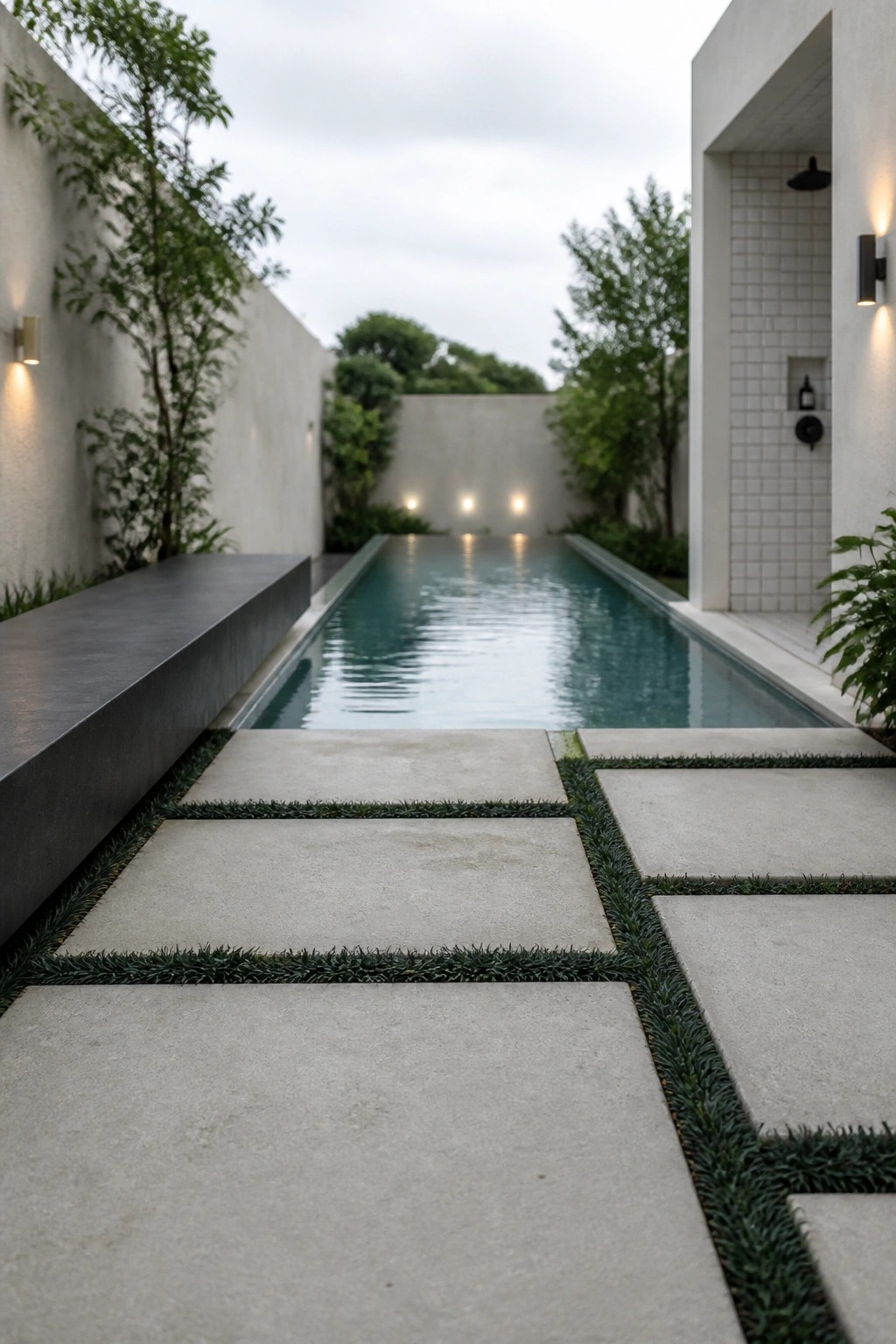 25 Elegant Pool Patio Layout Ideas That Improve Flow Around the Water A narrow rectangular pool with light gray concrete pavers separated by grass strips leading toward the water, flanked by white walls and trees.