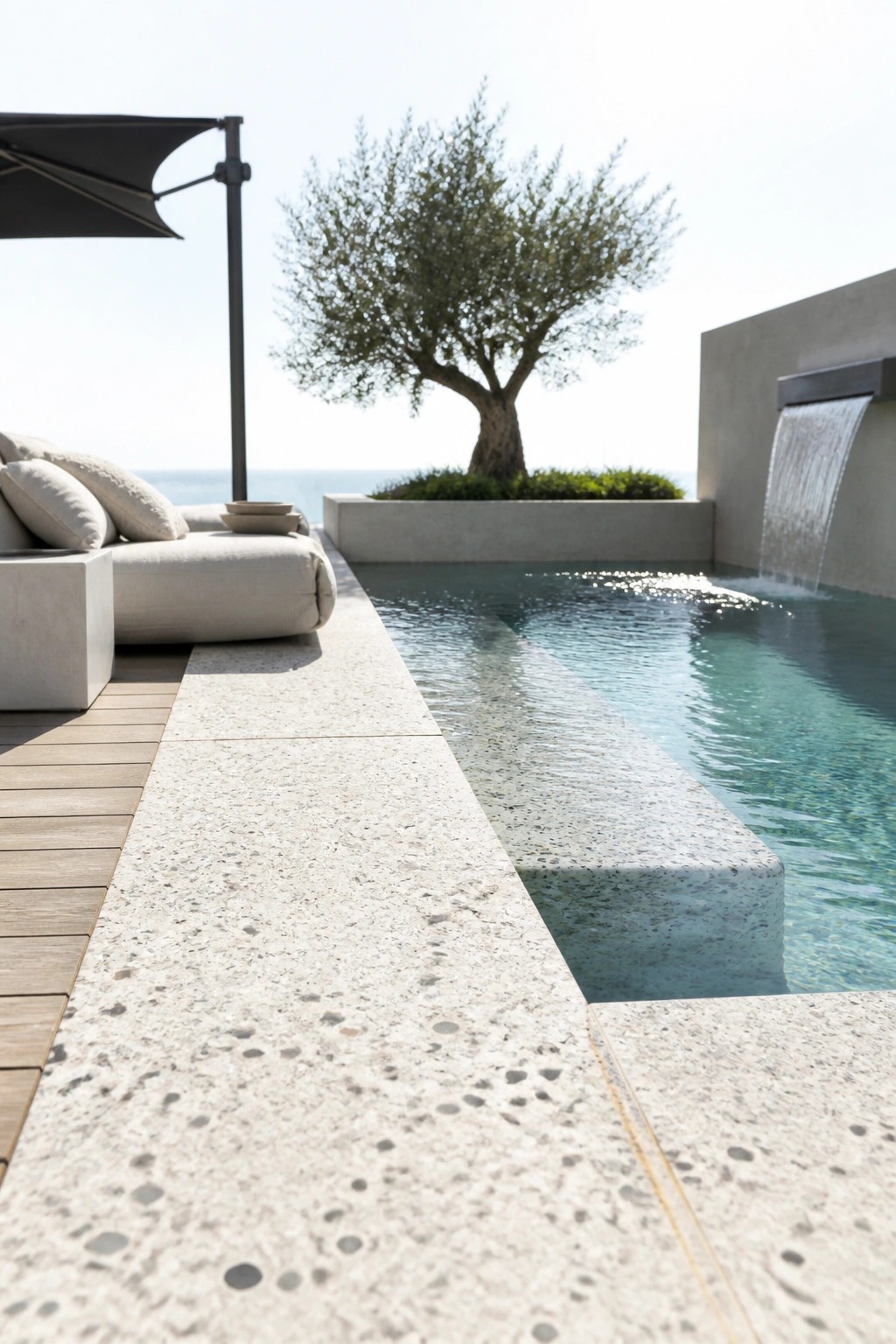 A narrow modern inground pool with a built-in concrete waterfall on the right wall, light terrazzo decking, and a single lounge chair on the left.