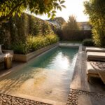 A narrow lap pool with a shallow sun shelf at one end, wooden deck with lounge chairs, built-in concrete bench with cushions, and surrounding plants and trees.