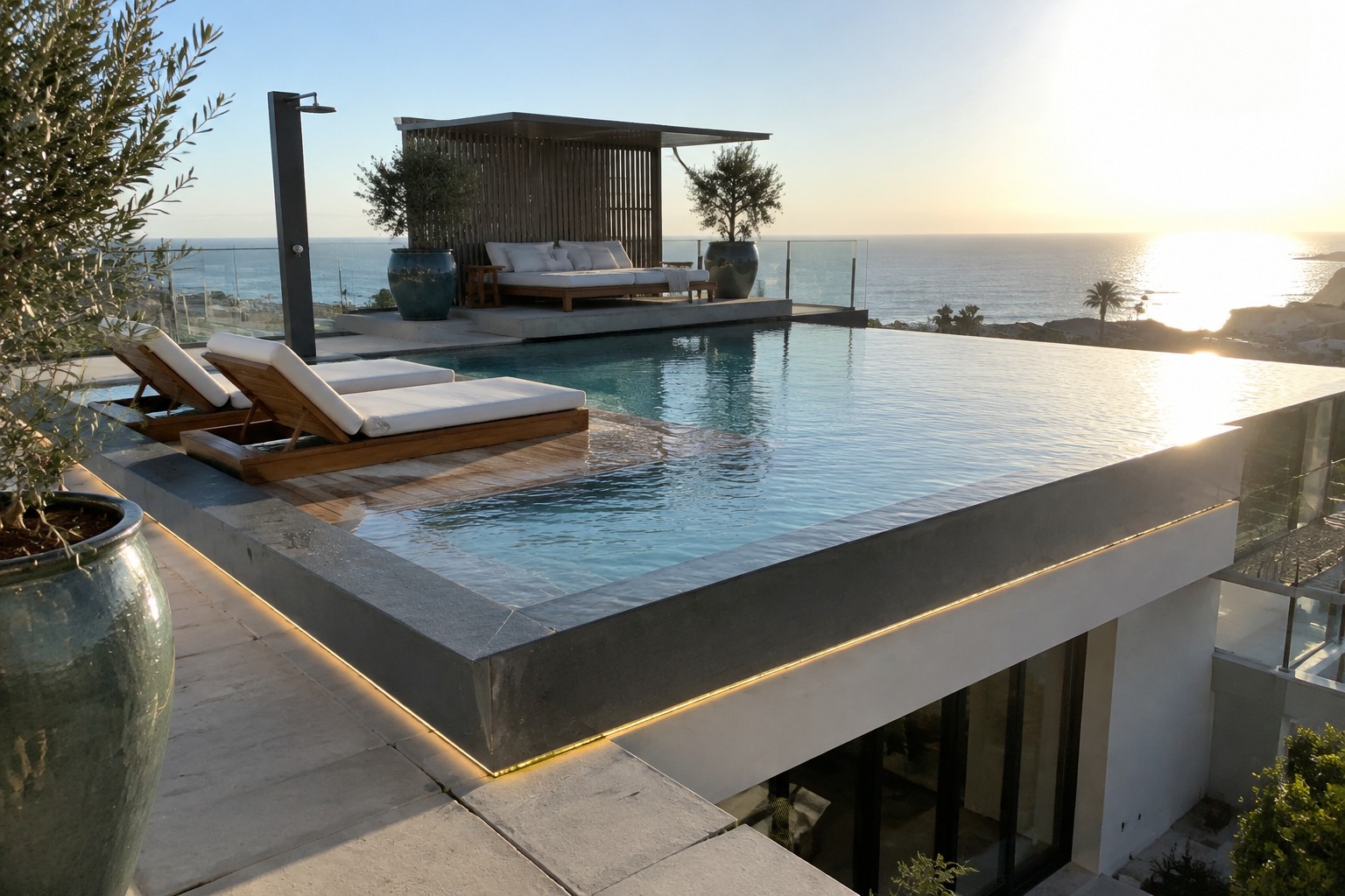 A modern infinity pool sits on a raised concrete terrace with wooden lounge chairs, a canopy daybed, and an ocean view at sunset.