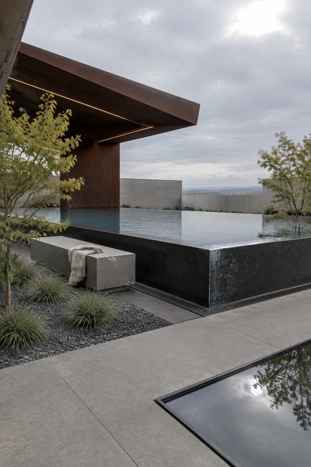 14 Elevated Infinity Pool Ideas Featuring Raised Architectural Structures Modern infinity pool with raised dark stone edge, concrete bench with throw, gravel bed, and young trees beside a rusted metal overhang.