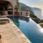 An infinity pool with light stone coping on a terraced patio, two lounge chairs, terracotta pots, and a view of the sea at sunset.