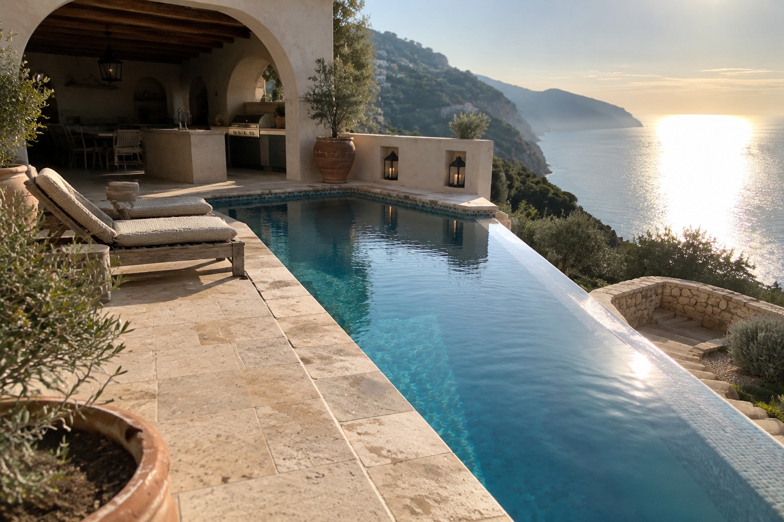 15 Mediterranean Infinity Pool Designs Using Stone and Architectural Details An infinity pool with light stone coping on a terraced patio, two lounge chairs, terracotta pots, and a view of the sea at sunset.