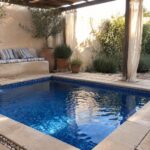 Tranquil blue-tiled pool in sunny courtyard with striped cushions, pergola, and terracotta pots
