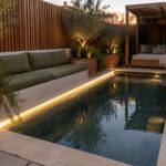 A narrow rectangular plunge pool with a continuous linear LED light strip set into the light stone coping, surrounded by potted olive trees and outdoor seating at dusk.