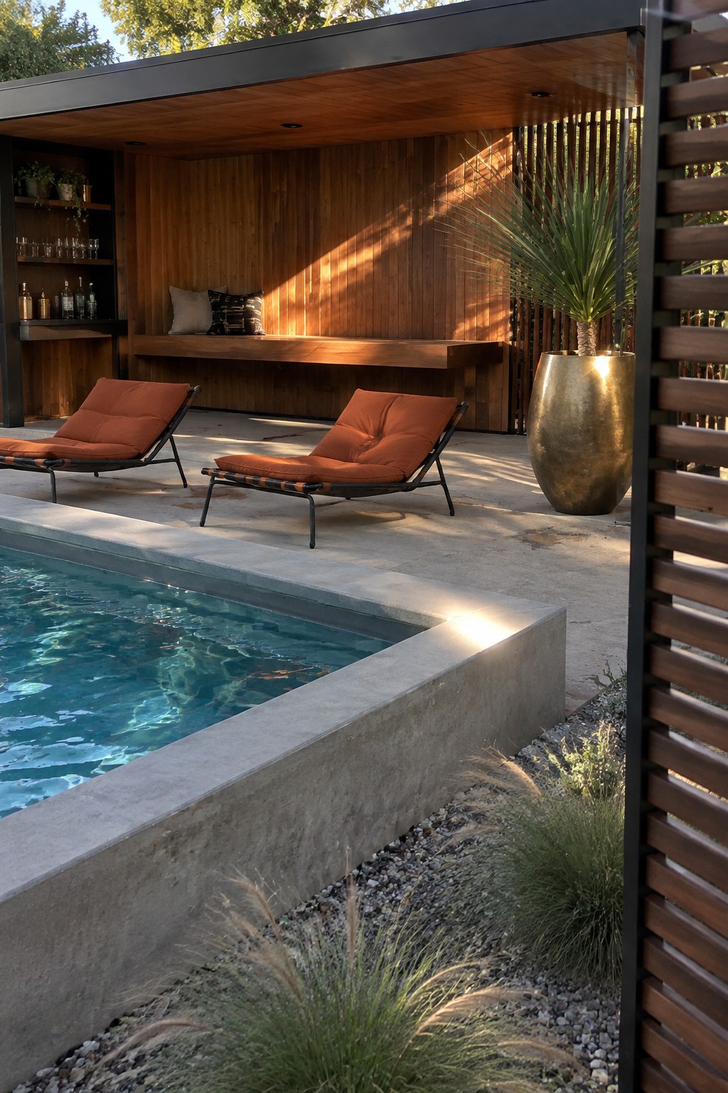 14 Functional Above Ground Pool Cabana Ideas for Shaded Lounging An outdoor pool area with two orange lounge chairs on concrete paving, a modern wooden cabana with built-in bench and bar shelves, and a large gold planter beside vertical wood screening.