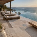 An outdoor patio with striped daybeds under a wooden pergola beside an infinity pool overlooking the ocean, with a linear fire pit at the pool edge.