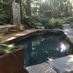 A natural plunge pool with dark water and pebble bottom is edged with irregular gray stone slabs, next to a wooden deck with bench and a stone outdoor shower in a wooded garden.