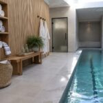 An indoor pool area with built-in wooden shelving holding stacks of rolled white towels, black wall hooks with hanging towels, a wooden bench, and a long lap pool with light blue water.