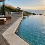 An infinity pool with light natural stone coping along the edge next to a wooden deck with lounge chairs, overlooking the ocean.