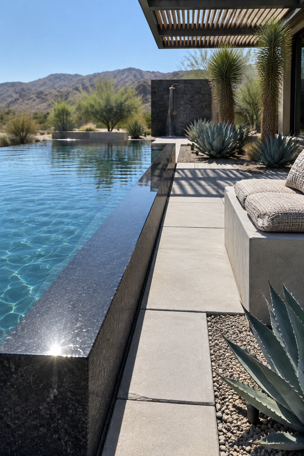 16 Sophisticated Infinity Pools With Natural Stone Edge Treatments An infinity pool with dark stone edging runs alongside light concrete pavers, with a concrete bench, gravel, and desert plants visible in the background.