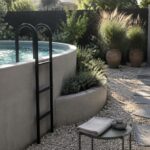A round concrete above-ground pool with a black ladder sits in a gravel yard next to large potted grasses, an outdoor shower on a concrete wall, and a path of gray stepping stones.